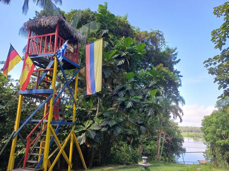 Una colorida torre de observación de madera con techo de paja se alza en un exuberante jardín selvático. Está decorada con banderas de Alemania, España, Grecia y Colombia. Al fondo, a la derecha, se ve una vista panorámica de un lago tranquilo bajo un cielo azul claro.