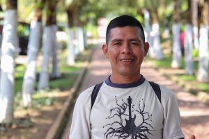 Retrato de medio cuerpo de un hombre adulto, con una expresión amable y una sonrisa ligera. Viste una camiseta de color crema con un diseño gráfico negro de una guitarra cuyas cuerdas se transforman en ramas de árboles. Se encuentra en un sendero peatonal al aire libre, rodeado de una hilera de troncos de árboles pintados de blanco que crean un efecto de profundidad. El fondo está suavemente desenfocado, resaltando la figura del hombre bajo una luz natural clara.