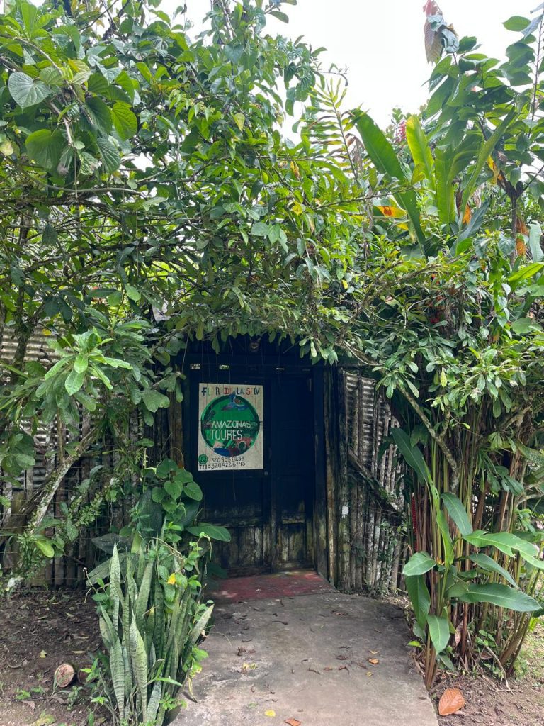 Entrada principal de Flor de la Selva Amazonas. Se observa una puerta de madera oscura enmarcada por abundante vegetación tropical. En la puerta cuelga un cartel con el logo de la agencia y números de contacto, rodeado de una estructura rústica de madera.