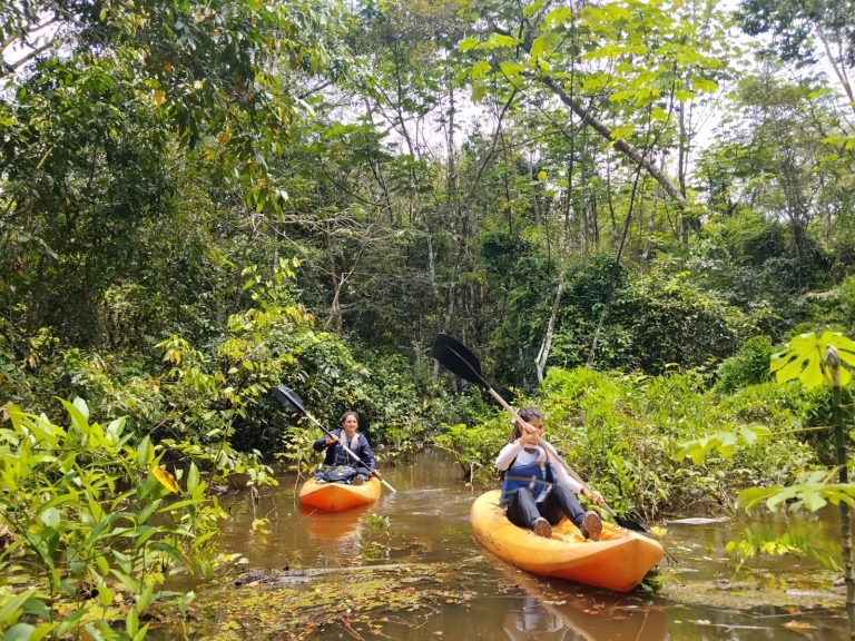 Dos personas navegando en kayaks individuales de color naranja por un canal estrecho de aguas tranquilas en la selva amazónica. La escena muestra una densa vegetación tropical de color verde intenso que envuelve el recorrido, destacando la experiencia de ecoturismo y aventura en la naturaleza.