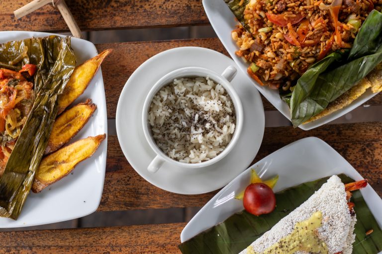 Vista cenital de cuatro platos de comida típica servidos sobre una mesa de madera. Se observa un tazón central con arroz blanco espolvoreado con semillas, un plato con pescado envuelto en hoja de plátano acompañado de tajadas de plátano frito, un plato de arroz con verduras y carne, y una preparación blanca tipo tapioquita decorada con una fruta pequeña. Los alimentos están presentados de forma artesanal y colorida.