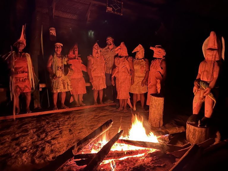 Escena nocturna en el interior de una maloca iluminada por una fogata en el suelo. Un grupo de personas, varias de ellas usando máscaras ceremoniales alargadas y trajes de corteza de árbol que representan seres mitológicos, posan junto a un hombre en ropa de campo. A la derecha, destaca una estatua tallada en madera de una figura antropomorfa. La luz de las llamas crea sombras dramáticas contra el suelo de tierra.