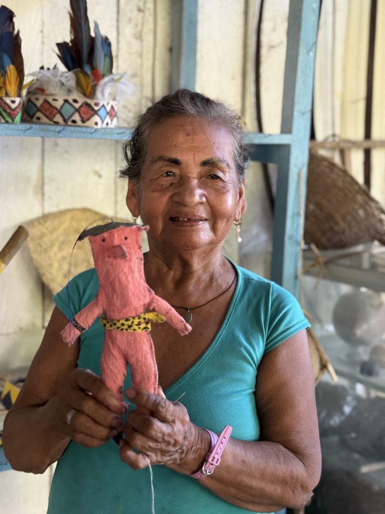 Retrato de una mujer anciana indígena Magüta - Tikuna con una sonrisa amable, vistiendo una camiseta verde azulada. Sostiene con ambas manos una pequeña figura artesanal de un delfín rosado antropomórfico, hecho de fibras naturales con un cinturón de jaguar. Al fondo, se observa un estante con otras artesanías, incluyendo una corona de plumas coloridas y cestos tejidos.