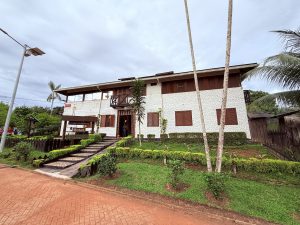 Vista frontal completa del 'Hotel Waira Selva' desde la calle. Se aprecia la edificación blanca de dos pisos con detalles en madera, el acceso por escaleras de piedra entre jardines y el pavimento de adoquines rojos en primer plano.