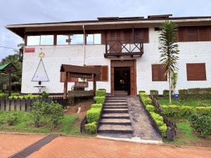 Fachada del Hotel Waira Selva en Puerto Nariño, Amazonas, Colombia. El edificio es de dos pisos con paredes blancas de textura rugosa, detalles en madera marrón y una rampa de acceso flanqueada por arbustos verdes.