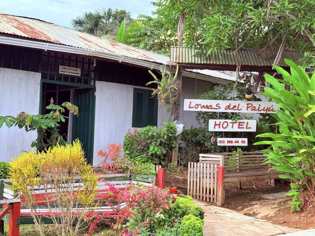 Fachada rústica del Hotel Lomas del Paiyú rodeada de jardines tropicales. Incluye letrero informativo con teléfono y bancas de madera en el acceso principal.
