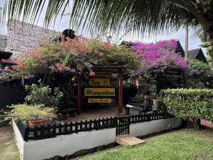 Detalle de la fachada del Restaurante Las Margaritas, destacando su jardín ornamental con flores tropicales y techo de paja tradicional al fondo. La entrada presenta un estilo rústico con madera oscura y señalización colorida.