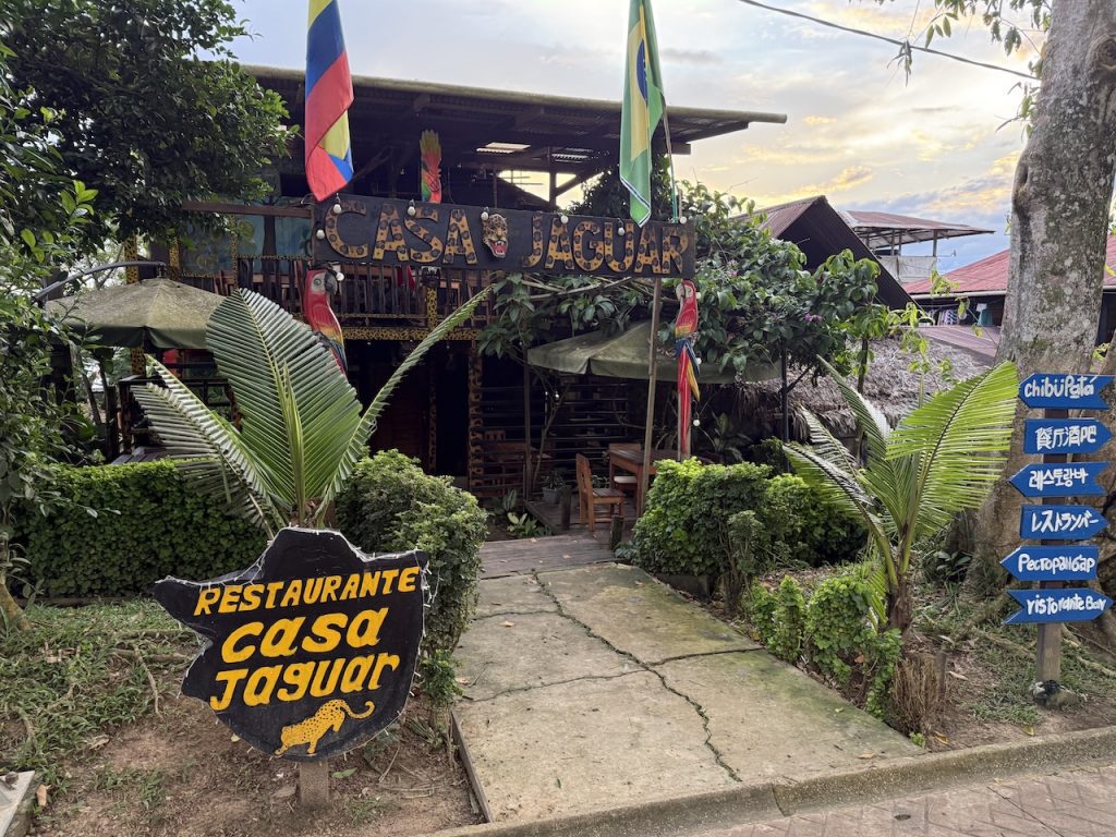 Vista ligeremanete ladeada de la fachada exterior rústica del restaurante 'Casa Jaguar', una estructura de madera de dos pisos rodeada de vegetación tropical. Cuenta con banderas de Colombia y Brasil, un letrero principal con estampado de jaguar y un poste de señalización con flechas azules en varios idiomas.