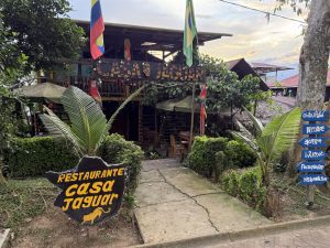 Vista ligeremanete ladeada de la fachada exterior rústica del restaurante 'Casa Jaguar', una estructura de madera de dos pisos rodeada de vegetación tropical. Cuenta con banderas de Colombia y Brasil, un letrero principal con estampado de jaguar y un poste de señalización con flechas azules en varios idiomas.