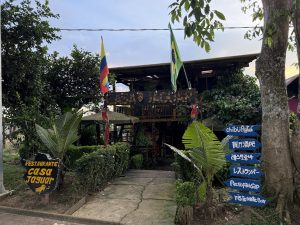 Fachada exterior rústica del restaurante 'Casa Jaguar', una estructura de madera de dos pisos rodeada de vegetación tropical. Cuenta con banderas de Colombia y Brasil, un letrero principal con estampado de jaguar y un poste de señalización con flechas azules en varios idiomas.