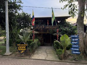 Vista frontal de la fachada exterior rústica del restaurante 'Casa Jaguar', una estructura de madera de dos pisos rodeada de vegetación tropical. Cuenta con banderas de Colombia y Brasil, un letrero principal con estampado de jaguar y un poste de señalización con flechas azules en varios idiomas.