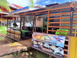 Entrada al Restaurante Los Amazónicos, un establecimiento de comida típica regional. Se aprecia el área de comedor al aire libre y un banner publicitario con imágenes de pescados, caldos y platos locales bajo un ambiente acogedor y rústico.
