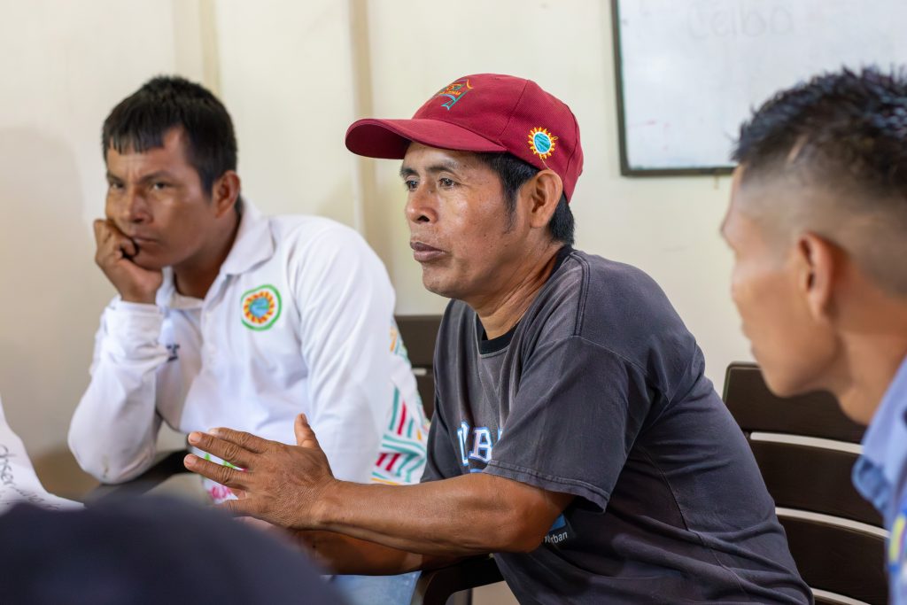 Un líder comunitario con gorra roja interviene durante una mesa de diálogo. Los asistentes muestran una actitud de escucha atenta, reflejando un proceso de toma de decisiones o intercambio de ideas en un entorno local.