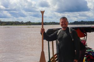 Retrato de un hombre de pie en una embarcación sobre el río Amazonas. El hombre, de complexión robusta y sonrisa cálida, viste una camiseta negra de manga larga y lleva binoculares colgados al cuello. Sostiene verticalmente un remo de madera tradicional cuya parte superior tiene una forma cóncava distintiva. Al fondo, se observa la inmensidad del río de aguas color café bajo un cielo nublado y una línea de selva distante.