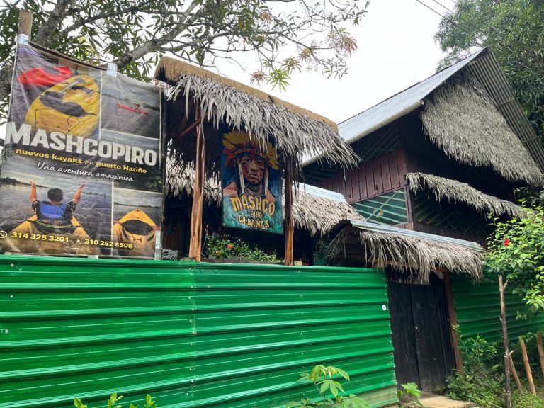Fachada de Mashco Piro Cabañas en Puerto Nariño, Amazonas. Se observa una construcción de madera con techos de paja tradicionales, un cartel publicitario de tours en kayak y una valla verde en primer plano rodeada de vegetación tropical.