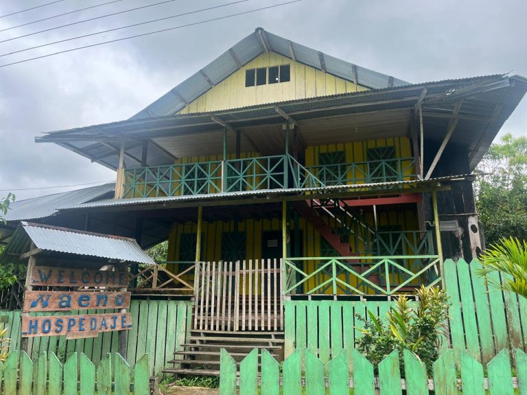 Instalaciones del Hospedaje NAEMW en la selva amazónica, mostrando una casa tradicional de madera pintada de amarillo y verde con balcón y cerca. Un cartel de bienvenida de madera rústica da la bienvenida a los huéspedes a un alojamiento auténtico y pintoresco en Puerto Nariño.