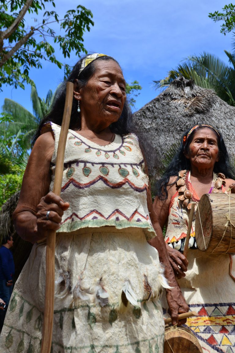 Fotografía de ángulo bajo de dos mujeres indígenas mayores vestidas con trajes tradicionales de fibra natural decorados con patrones geométricos y plumas. La mujer en primer plano sostiene un bastón de mando de madera, mientras que la mujer al fondo carga un tambor artesanal. Ambas lucen bandas ceremoniales en la cabeza bajo un cielo azul brillante y frente a una maloca de paja.