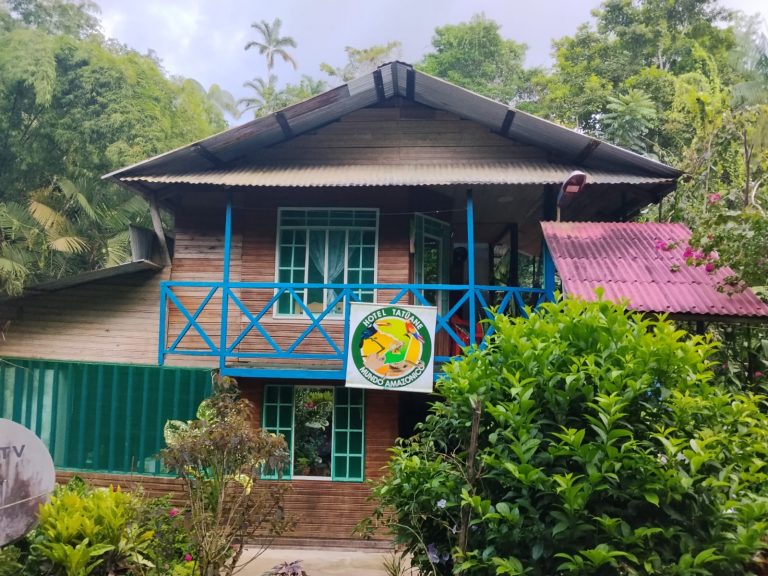 Fachada del Hotel Tatuane - Mundo Amazónico. Una casa de madera de dos niveles con balcones azules, ventanales verdes y un cartel circular con el logo del hotel. La construcción está rodeada de exuberante vegetación selvática, árboles altos y plantas ornamentales bajo un cielo nublado.