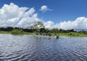 Una lancha motorizada blanca navega por un río amplio bajo un cielo azul brillante con grandes nubes blancas acumuladas. En la embarcación viaja un grupo de personas, varias de ellas sosteniendo cámaras con lentes largos. El río está rodeado de vegetación baja y selva densa al fondo.