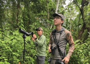 Dos hombres realizan avistamiento de aves en una selva densa. El hombre en primer plano, con gafas y binoculares colgando del cuello, mira hacia arriba con atención. Detrás de él, un guía local ajusta un telescopio profesional montado sobre un trípode. Ambos están rodeados de abundante vegetación verde y visten ropa técnica de campo.