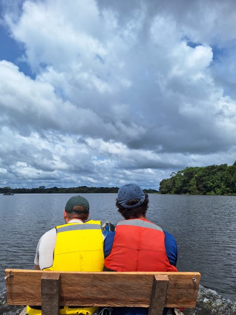 Fotografía tomada desde la parte trasera de una lancha simple con un banco de madera rústica en primer plano. Dos hombres están sentados de espaldas; el de la izquierda lleva una gorra verde y chaleco amarillo, el de la derecha gorra azul y chaleco naranja. Miran hacia un río ancho y tranquilo bordeado por densa vegetación selvática a lo lejos. Un barco muy pequeño se ve a la izquierda. El cielo está lleno de nubes cúmulos grandes y grises, con parches de azul.