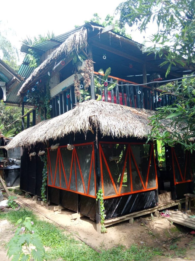 Fachada exterior de un ecolodge rústico y sostenible en la selva amazónica. Cabaña de dos niveles con techo de paja, balcón abierto decorado y una planta baja con diseño moderno y vibrantes toques naranjas, ofreciendo una experiencia auténtica y natural en Puerto Nariño.