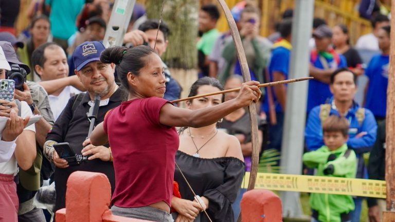 Mujer indígena practicando tiro con arco tradicional durante un evento cultural en el Amazonas. La joven sostiene un arco de madera y flecha con precisión, rodeada de espectadores y fotógrafos en un entorno festivo.