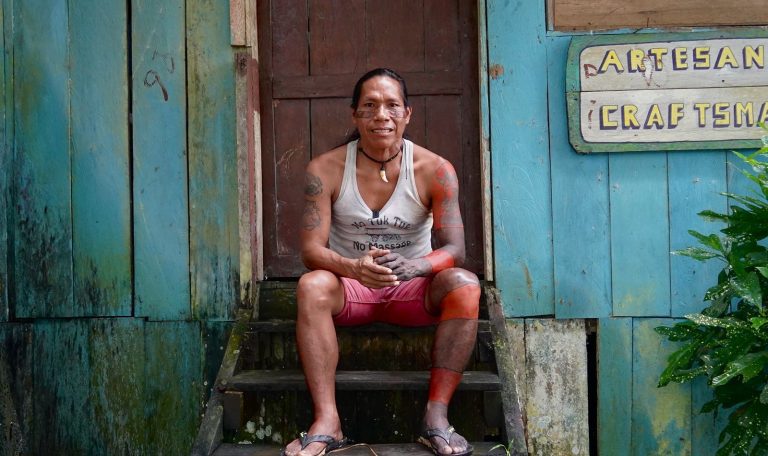 Retrato de un hombre indígena con pinturas corporales tradicionales sentado en la entrada de un taller de artesanías en Puerto Nariño, Amazonas. Detrás, una pared de madera azul con el letrero "Artesanías / Craftsman".