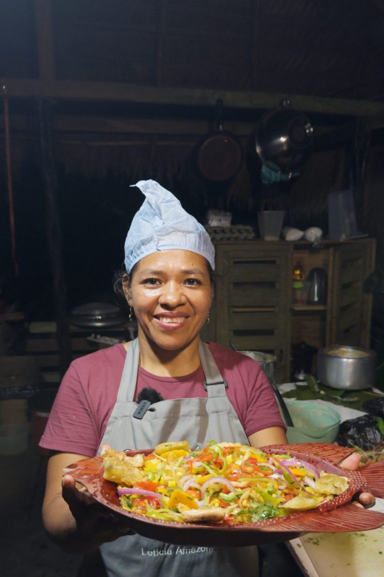 Cocinera local del Amazonas presentando una fuente de verduras vibrante y colorido, servido en una fuente de madera tallada artesanalmente en forma de pescado, en un entorno culinario tradicional de Puerto Nariño, Amazonas.