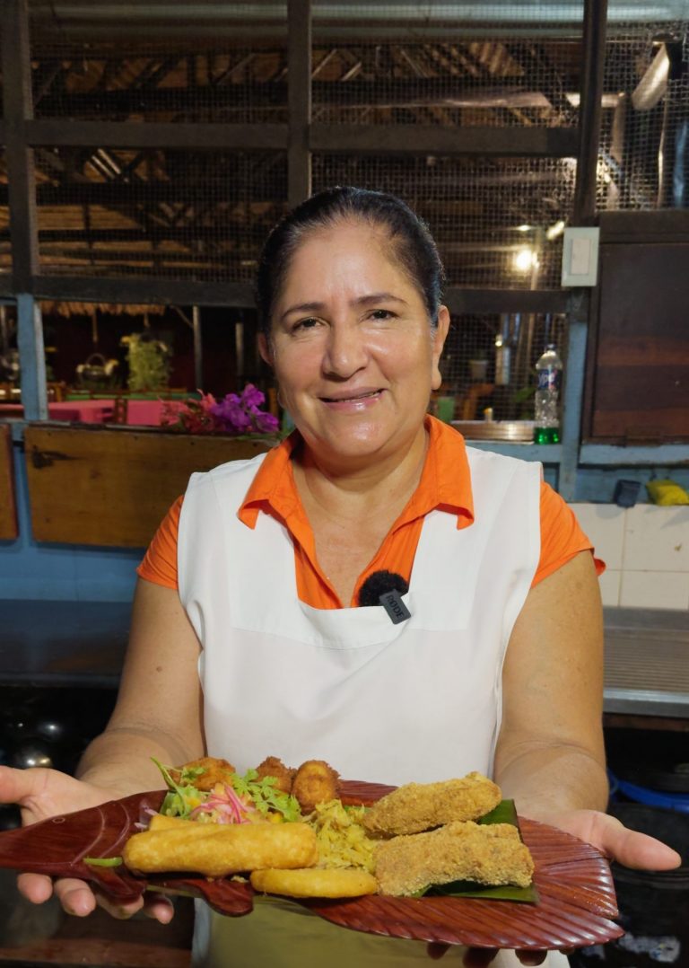 Cocinera local mostrando una bandeja de madera con especialidades fritas, incluyendo lo que parecen ser croquetas y empanadillas, servidas en un ambiente de cocina rústica.