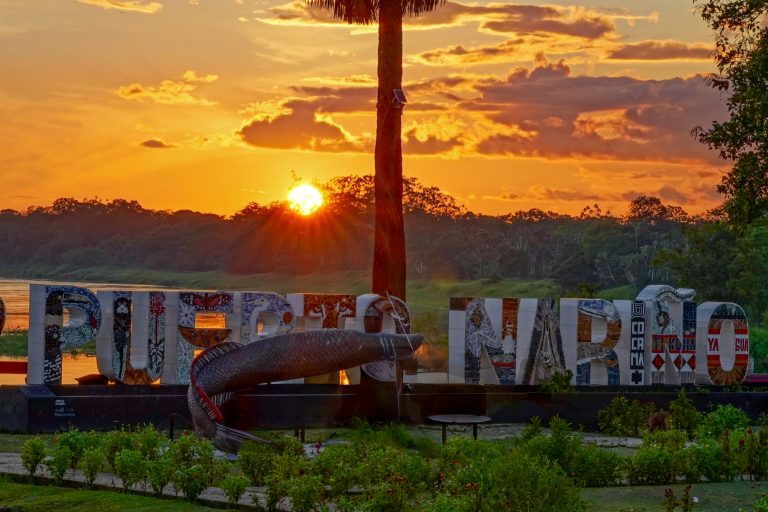 Fotografía de un letrero monumental con el nombre 'PUERTO NARIÑO' decorado con mosaicos coloridos que representan la fauna y flora local. Frente a las letras, se encuentra una gran escultura de un pez pirarucú. La escena ocurre durante un atardecer vibrante, con el sol bajo proyectando rayos anaranjados detrás de una silueta de selva y un río tranquilo. En primer plano, hay un jardín cuidado con arbustos bajos.