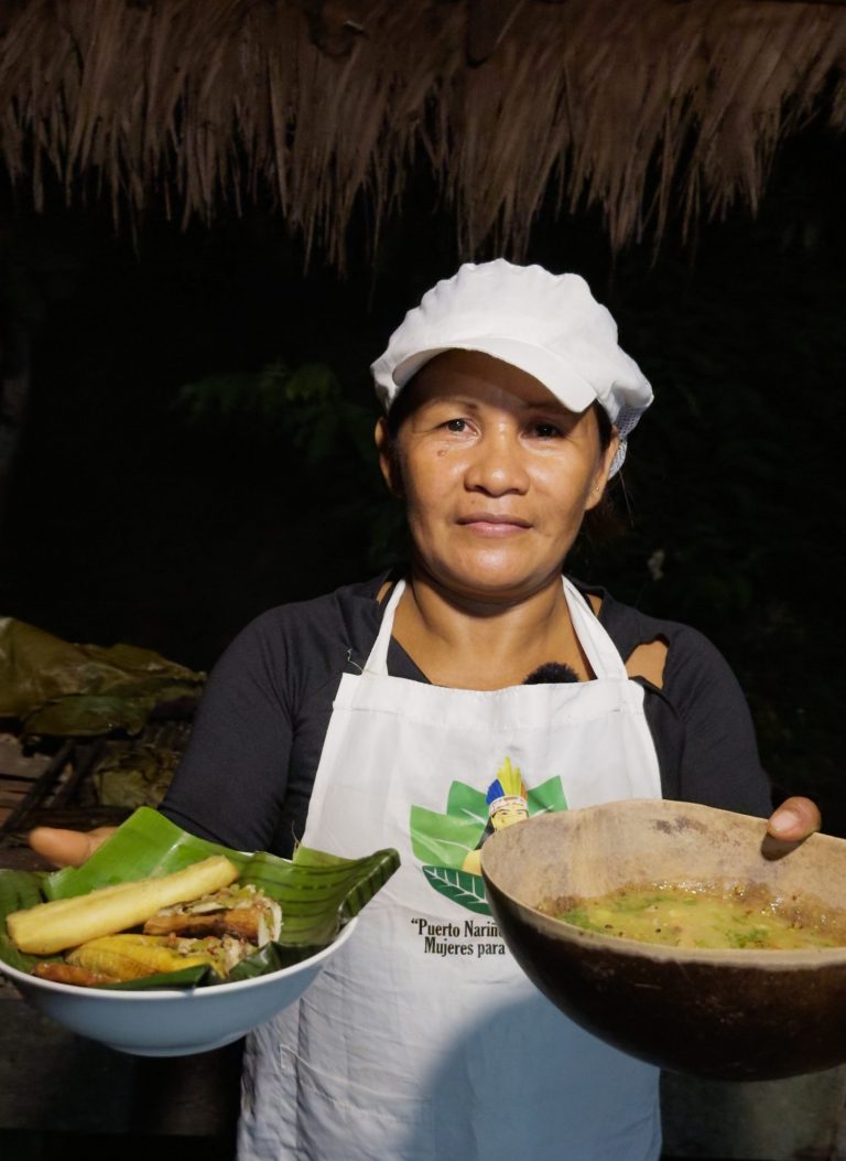 Cocinera local presentando un menú tradicional amazónico servido en un plato de loza y un recipiente de totumo o madera, destacando el uso de ingredientes locales y recipientes artesanales.