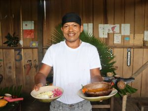 Retrato de un hombre indígena Magüta sonriendo mientras sostiene dos cuencos artesanales con comida tradicional. En su mano derecha presenta un plato con fariña y flores rosadas decorativas, y en la izquierda, un cuenco de madera con un caldo o salsa espesa de color naranja. Viste una camiseta blanca y un pañuelo negro en la cabeza. El fondo es una pared de madera decorada con dibujos, pintura y hojas de palma.