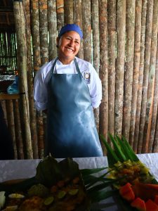 Retrato de una mujer cocinera sonriendo cálidamente frente a una pared de troncos de madera verticales. Viste un uniforme de chef blanco, un delantal largo de color azul grisáceo y una gorra de cocina azul. En primer plano, sobre una mesa con mantel blanco, se aprecian platos decorados con hojas de palma, carambolas y otros ingredientes regionales.