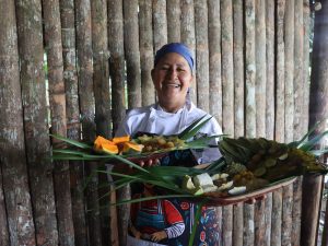 Retrato de una chef sonriendo ampliamente frente a una pared de troncos de madera. Viste un uniforme blanco de cocina, un gorro azul y un delantal decorado con una ilustración colorida. Sostiene con ambas manos dos bandejas de madera cargadas de comida tradicional. Los platos incluyen pescados envueltos en hojas de bijao, arroz, huevo cocido, aguacate, trozos de papaya y carambolas, todo decorado con largas hojas verdes que sobresalen de las bandejas.