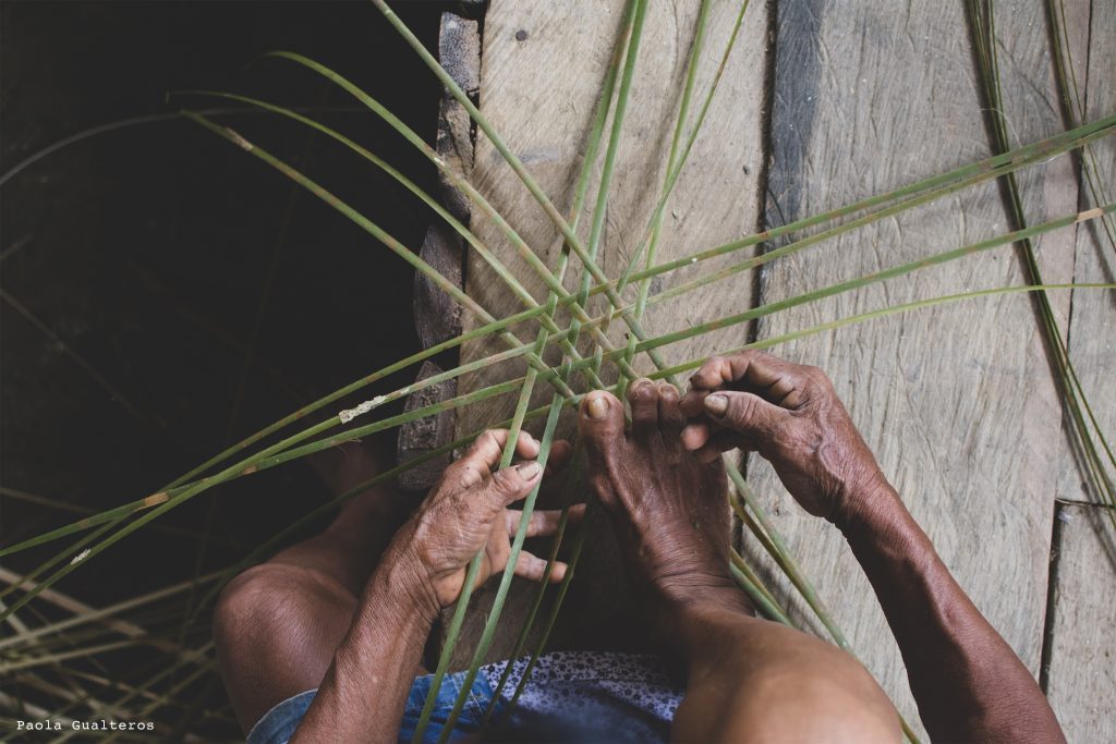 Vista cenital de una artesana indígena en Puerto Nariño utilizando sus manos y pies para tejer una canasta tradicional con fibras naturales verdes. Primer plano documental de la técnica de cestería amazónica en un piso de madera rústico.